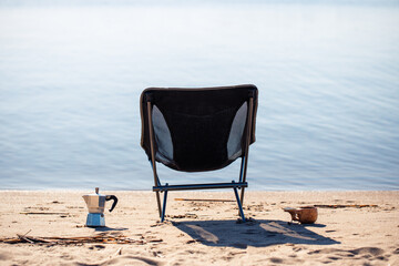 Fototapeta na wymiar Camping chair, coffee maker and mug in the morning on the seashore close-up background is blurred.