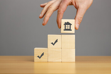 Hand putting wooden block pyramid with house and marks icon on wooden table grey background. Construction, engineering concept.