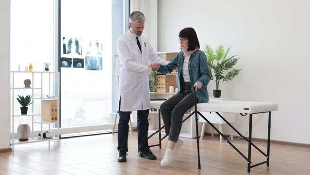 Confident male in lab coat assisting beautiful woman in neck brace with finding balance when walking with cane in clinic. Physiotherapist giving support while lady making first steps after injury.