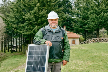 Middle aged bearded technician with voltaic solar panel near house in rural zone. Stand-alone...