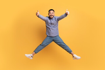 Fototapeta na wymiar Joyful Asian Man Jumping In Air And Showing Thumbs Up At Camera