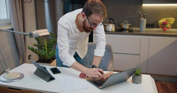 Top view young man working on project on laptop, standing at shared table in modern office kitchen. Businessman holding documents, involved in financial paperwork using computer e-banking application.