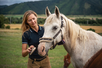 Young woman cleaning white gray horse with brush, her head next to animal