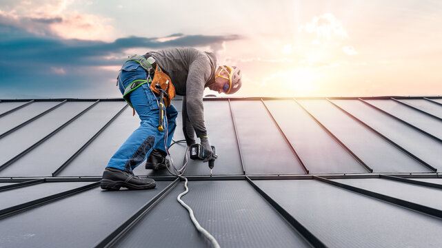 Work at heights. A tinman with a climbing harness and the tool belt on the roof is working with a cordless drill on sheeting.