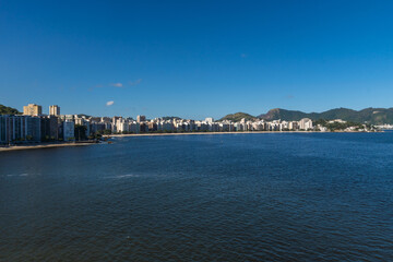 Fototapeta premium View of the edge of Praia de Icaraí, a neighborhood in Niterói, Rio de Janeiro, Brazil. Buildings and houses in front of the beach, surrounded by hills and nature. Bathed by Guanabara Bay. Sunny day
