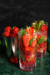 Glasses of fresh strawberry mojito and shaker on grunge green table against black wall