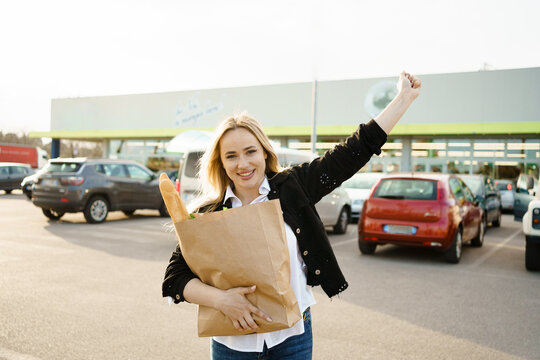An outdoor portrait capturing the joy of a smiling woman holding a paper bag full of fresh and healthy groceries outside a supermarket.