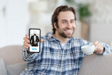 Smiling young caucasian man with stubble sits on sofa with coffee cup, shows phone with suit store on screen