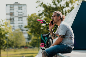 A middle-aged tattooed urban bmx bike rider is sitting in a skate park and calling on the phone.