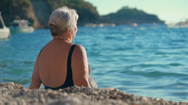 Elderly senior woman with gray hair, sitting in shallow calm sea near beach enjoying sunshine, view from behind
