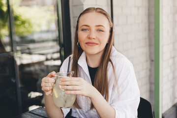 Attractive young woman with a glass of lemonade on a hot summer day. Beautiful female look dreaming and happy, she is smiling. Summer mood, 
fresh drinks. Girl sitting in cafe outside with lemonade.