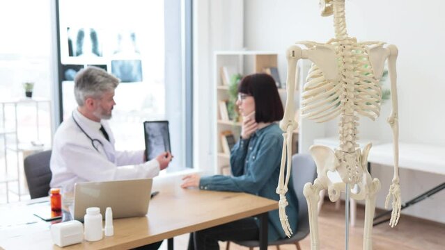 Focus on back view of teaching model located in consulting room of private practice with adults talking in background. Experts using skeleton as representation of human anatomy for patient education.