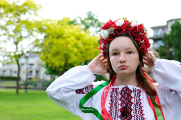 beautiful Strong brave Ukrainian young woman in red wreath of flowers of embroidered shirt with ribbons in hair stands against background of green trees looks into distance seriously power Glory