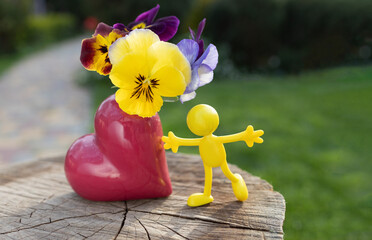Miniature bright toy figurine of a yellow man near a heart-shaped vase with pansies. concept of love, joy, happiness. positive thinking. flower gift. fun childhood