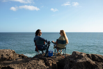 Happy Young Couple Sitting In Camping Chairs On Beach Rocks Near Ocean