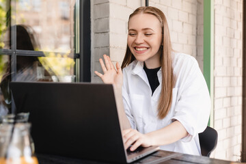 Close up of a beautiful young blonde woman at the cafe outdoors, having video call on laptop. Woman look at laptop camera and wave hand, hi, hello, welcome.