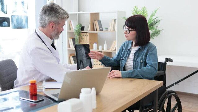 Confident adult in doctor's coat holding tablet while pretty lady in glasses sitting in wheelchair at writing desk in hospital. Efficient traumathologist analyzing CT scan image with patient at work.