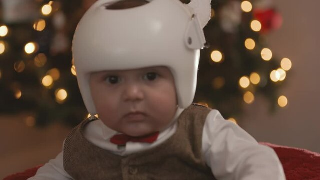 8 Month Old Baby Wearing Therapy Helmet Christmas Pose Turn to Camera. slow motion christmas pose of an 8 month old sitting in front of christmas tree wearing a cranial therapy helmet and turns toward