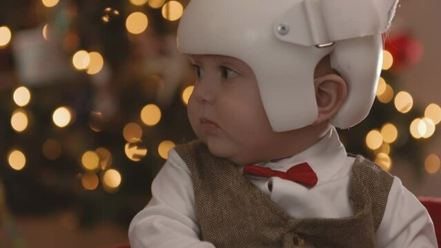 8 Month Old Baby Wearing Therapy Helmet Christmas Pose Tilt Up Turn to Camera. slow motion christmas pose of an 8 month old sitting in front of christmas tree wearing a cranial therapy helmet tilt up 