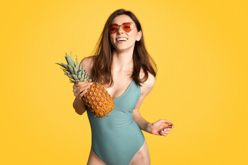 Ecstatic young woman model in one-piece swimsuit holding exotic fruit, one ripe pinapple and laughing, yellow background