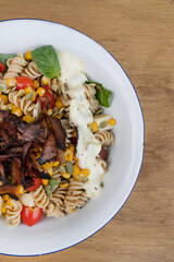 Top view of a Fusilli pasta salad with basil, crispy bacon, corn, cheese, tomato and a poached egg, in a white bowl on the wooden restaurant table.