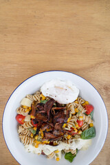 Top view of a Fusilli pasta salad with basil, crispy bacon, corn, cheese, tomato and a poached egg, in a white bowl on the wooden restaurant table.