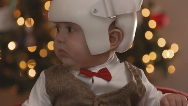 8 Month Old Baby Wearing Therapy Helmet Christmas Pose Tilt Up Look Left. slow motion christmas pose of an 8 month old sitting in front of christmas tree wearing a cranial therapy helmet tilt up