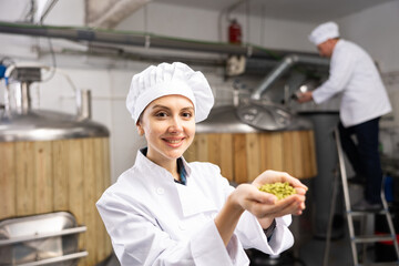 Portrait of positive woman brewmaster holding bunch of hops pellets in beer factory.