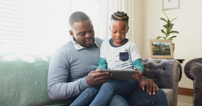 Happy african american father sitting on sofa with son on lap using tablet, in slow motion