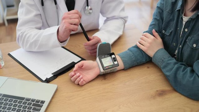 Close up view of bespectacled woman wearing wrist-type device while sitting at desk with physician in white coat. General practitioner estimating measurements before saving in patient record.