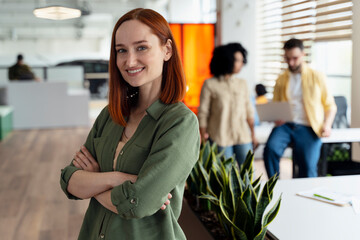Smiling confident young ginger woman manager with arms folded in corporate office building interior - obrazy, fototapety, plakaty