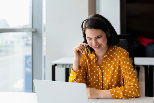 Pleasant young businesswoman, sales manager, executive director or recruiter in audio headset, sitting at table, using laptop having online meeting with partners or job interviews in office interior