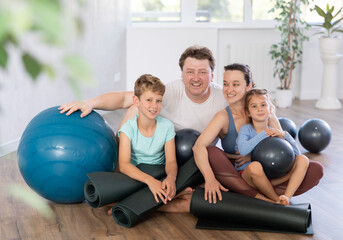 Portrait of happy family engaged in pilates sitting on the floor with black mats and softballs in hands in light room