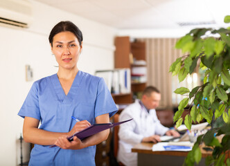 Portrait of polite Asian female doctor wearing blue uniform meeting patient in clinic, filling out medical form at clipboard - obrazy, fototapety, plakaty
