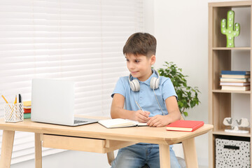 Boy writing in notepad near laptop at desk in room. Home workplace