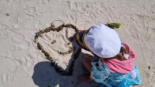 little girl with captain hat playing with white sand on a beach, drawing big smile heart emotion, view from the head. Creativity, holidays, childhood. High quality FullHD footage