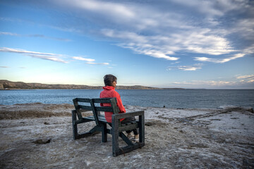 Senior woman contemplates the sea sitting on a bench on the rocks on a partly cloudy day, in Puerto Pirámides, Chubut, Argentina.