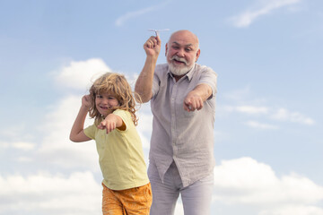 Old grandfather and young child grandson playing with toy paper plane against summer sky background.