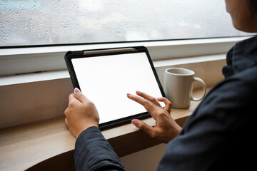 A hand holding a black tablet with a blank white screen on the window with a cup of black latte coffee and sitting on a wooden chair