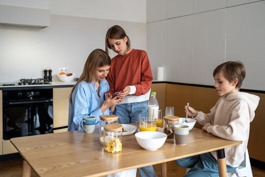 Teen daughter showing new video for social media on smartphone to mom during family breakfast on kitchen. Mother son eating sitting at table. Motherhood, parenting, rising children, mutual support. - Powered by Adobe