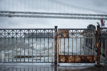 Stormy weather in San Francisco. In the foreground, a rusty gate is closed and locked at a boardwalk along the ocean.  In the background, crashing waves batter the railing.