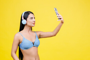 Portrait of young pretty girl with wireless headphones on head, taking selfie for social networks, smiling and laughing at phone camera, isolated on yellow background