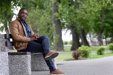 Obraz premium Stylish young freelancer in jeans and a brown bomber jacket relaxes in the fresh air in the park on a bench. Dark-skinned man inhales fresh air while walking in the park on a day off from work