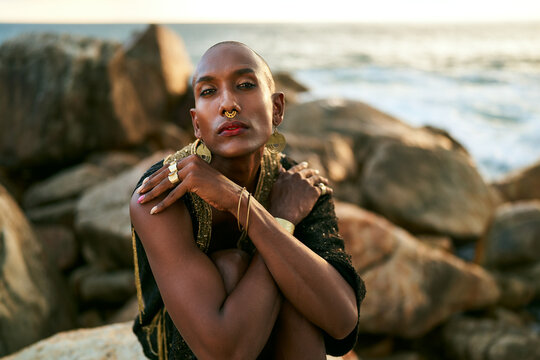 Epatage lgbtq black man poses on scenic ocean beach looks at camera demonstrates jewellery . Androgynous ethnic fashion model in posh dress looks at camera, sits on stone in