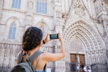 Young brunette with backpack taking picture of Barcelona cathedral with an smart phone. While exploring streets of historic city in daytime