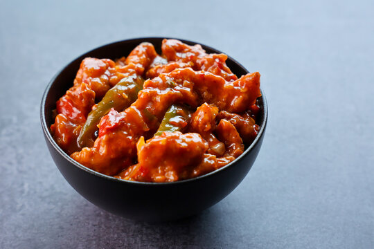 Schezwan Chicken or Dragon Chicken in black bowl at dark slate background. Szechuan Chicken is popular indo-chinese spicy dish with chilli peppers chicken and vegetables.