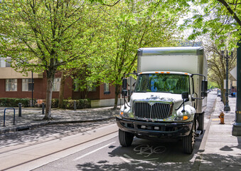 Obraz premium In demand white middle duty rig semi truck with day cab and box trailer for local delivery standing on the urban city street with trees shadow