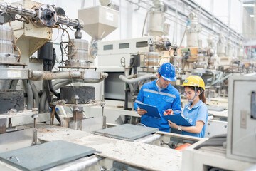 American male engineer and Asian female manager View the list of orders In a plastic industry factory wearing a uniform, holding a listnote and a helmet, there is a machine working.