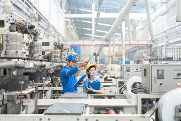 American male engineer and Asian female manager They are consulting together, wearing uniforms, radios, listnotes and helmets, machines working. in the plastic industry business plant