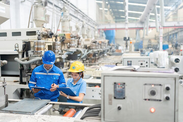 American male engineer and Asian female manager Looking down at plans in listnotes, wearing uniforms, radios and helmets, in a plastic industry factory. There is a working machine